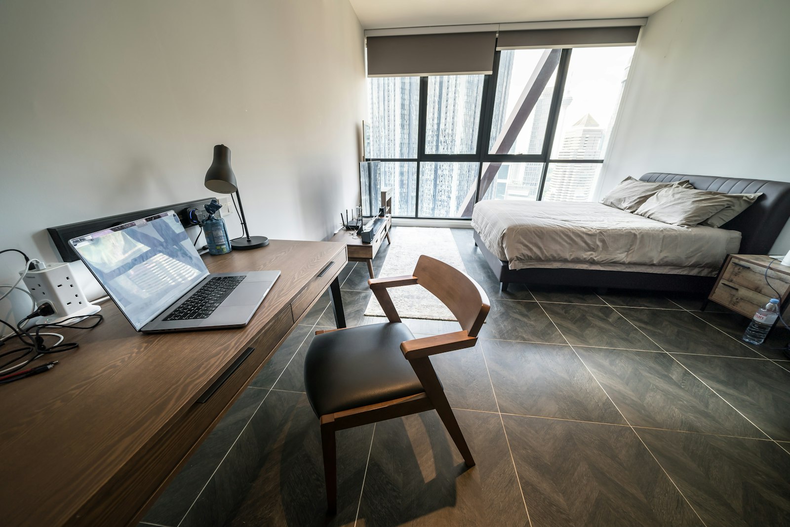 A long wooden desk with a laptop along one wall of a studio-style bedroom with a bed and a large window