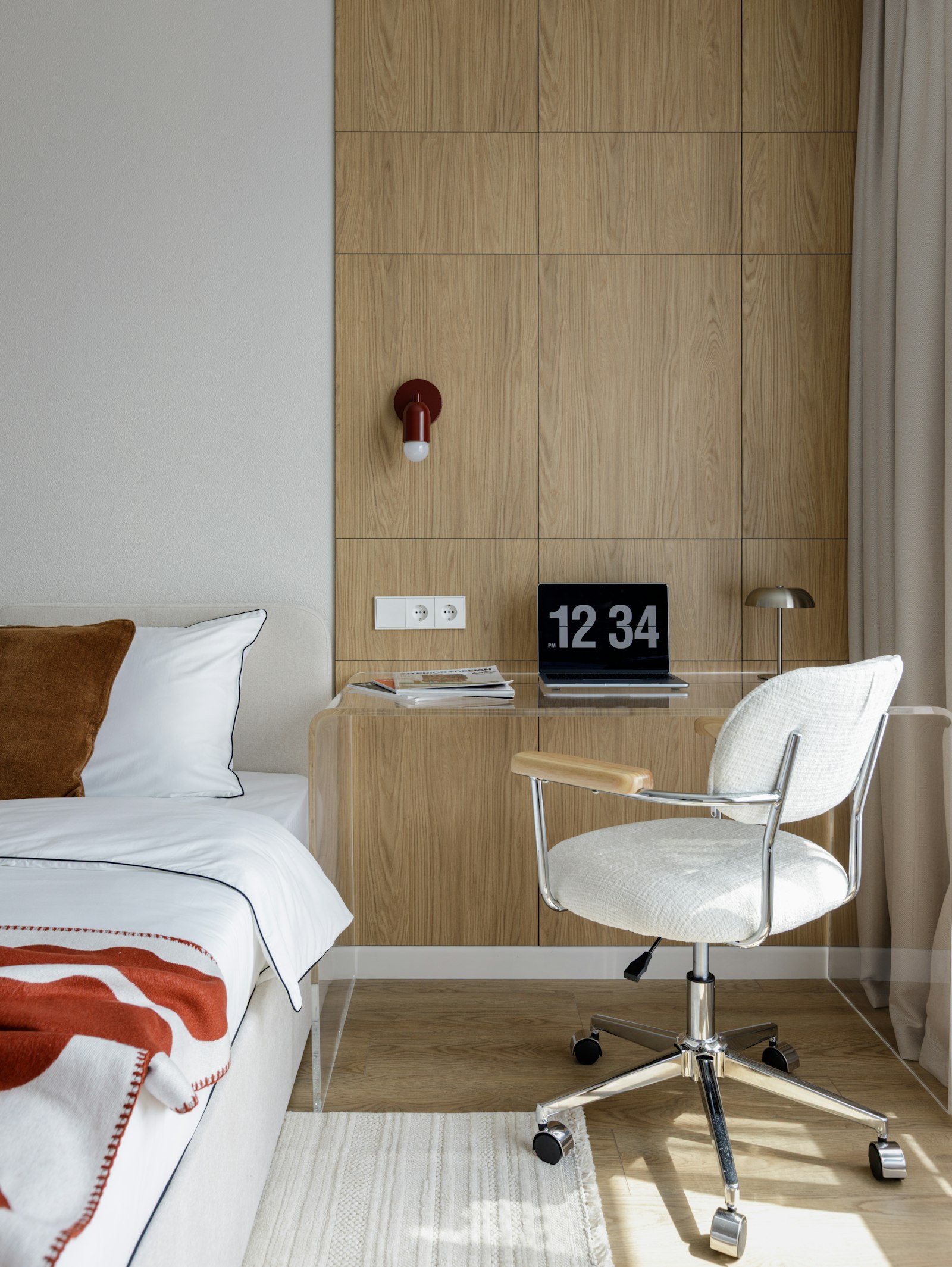 A clear acrylic desk with a laptop and an upholstered office chair next to a bed in a small bedroom