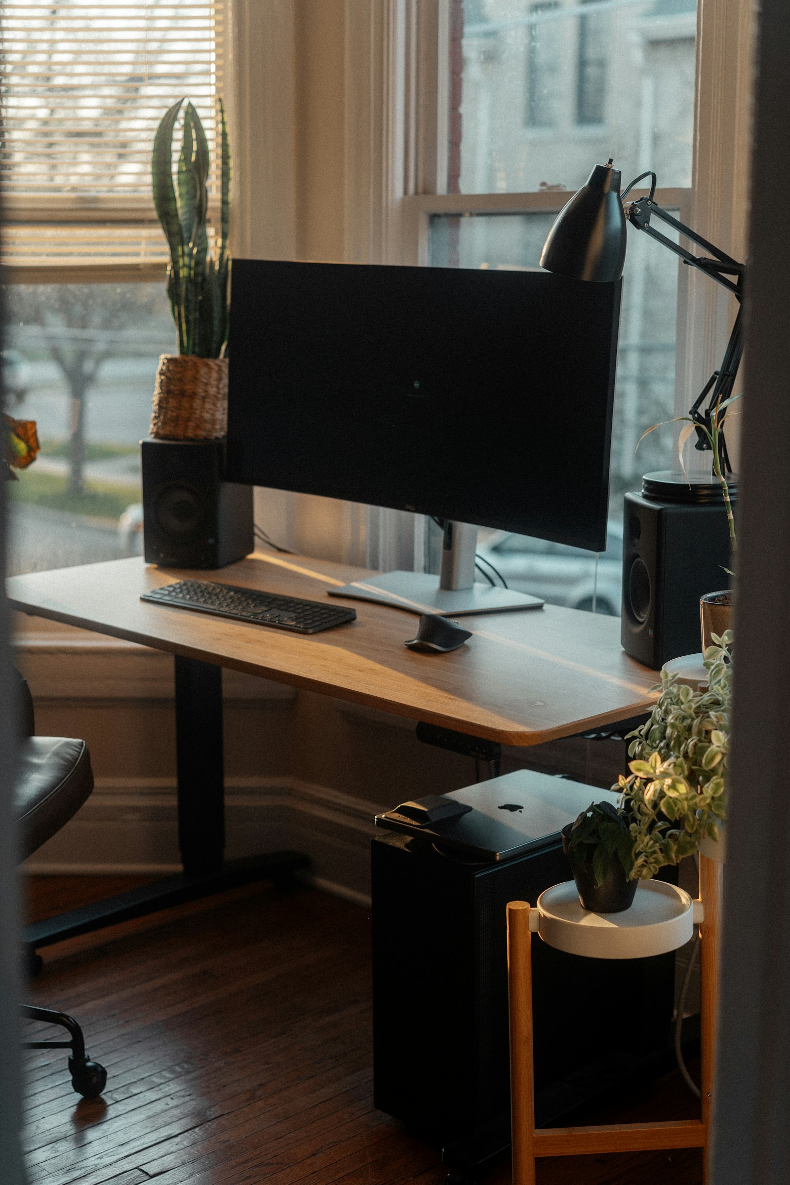A wooden desk by a window with a curved monitor, keyboard, speakers and plants